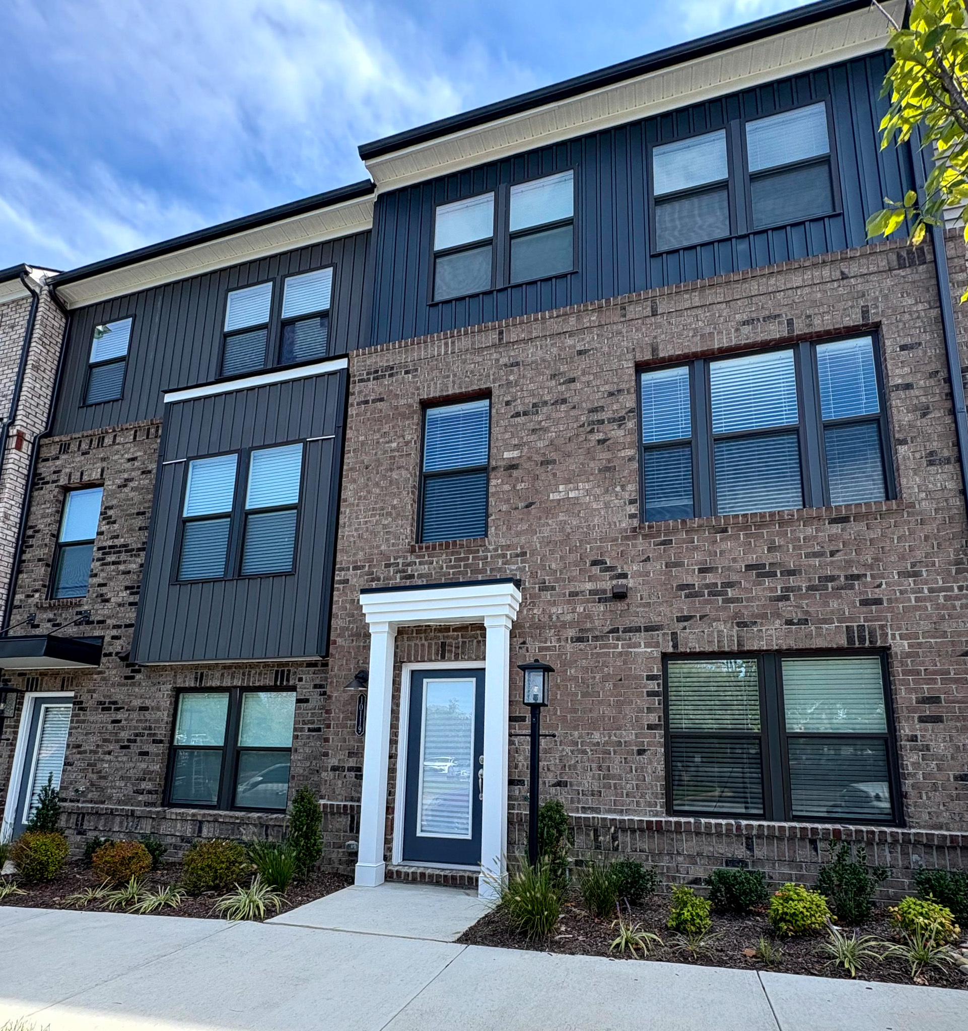 A modern three‑story townhouse with dark brick, dark siding panels, white trim around the front door, large windows, and neatly landscaped shrubs along the sidewalk belonging to Sponsored Residential Providers Tania and LeSean Matthews in Glen Allen, Virginia.