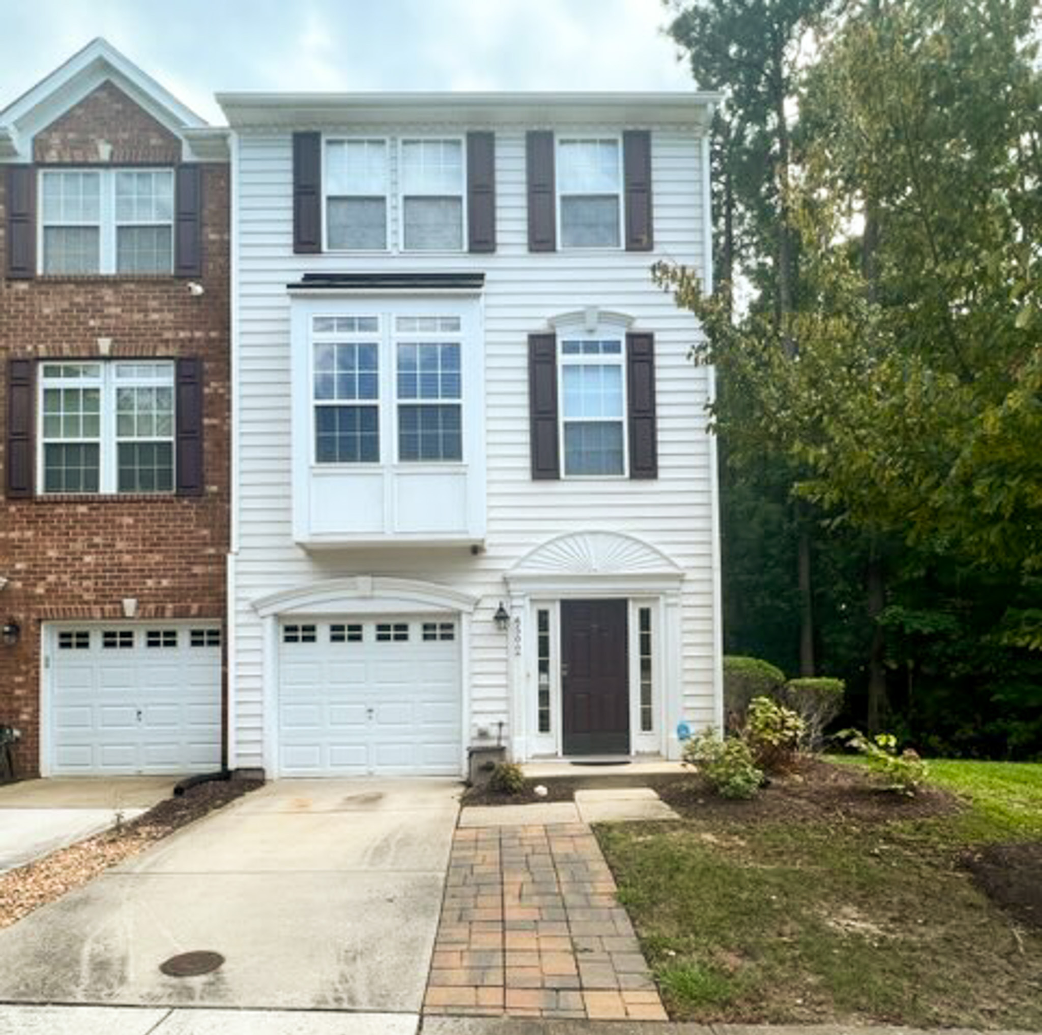 Three‑story townhome with white siding and brick exterior, single‑car garage, and front walkway in a cul‑de‑sac setting belonging to Sponsored Residential Provider Monika Soto in Williamsburg, VA