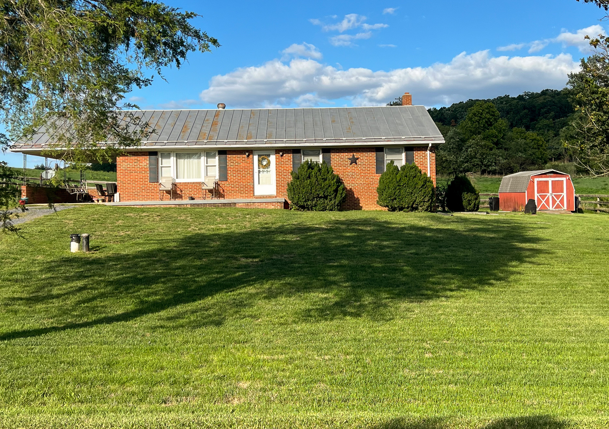 Brick ranch‑style house with white shutters, a large front lawn, shrubs along the facade, and a red barn in the background under a blue sky belonging to Sponsored Residential Provider Rebecca Sherfey in Edinburg, Virginia.