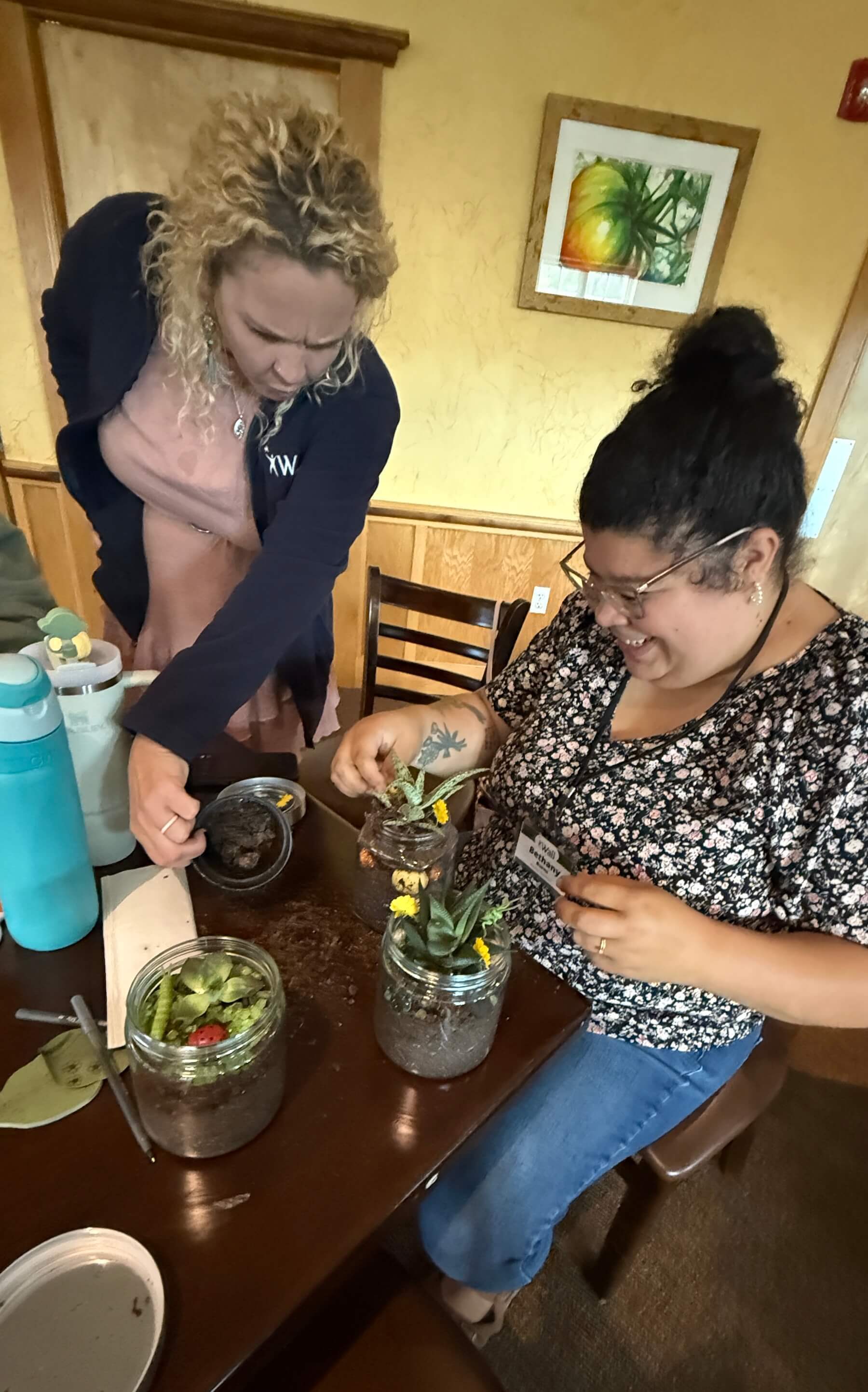 A woman sitting at a table, smiling and looking at a small plant in a jar
