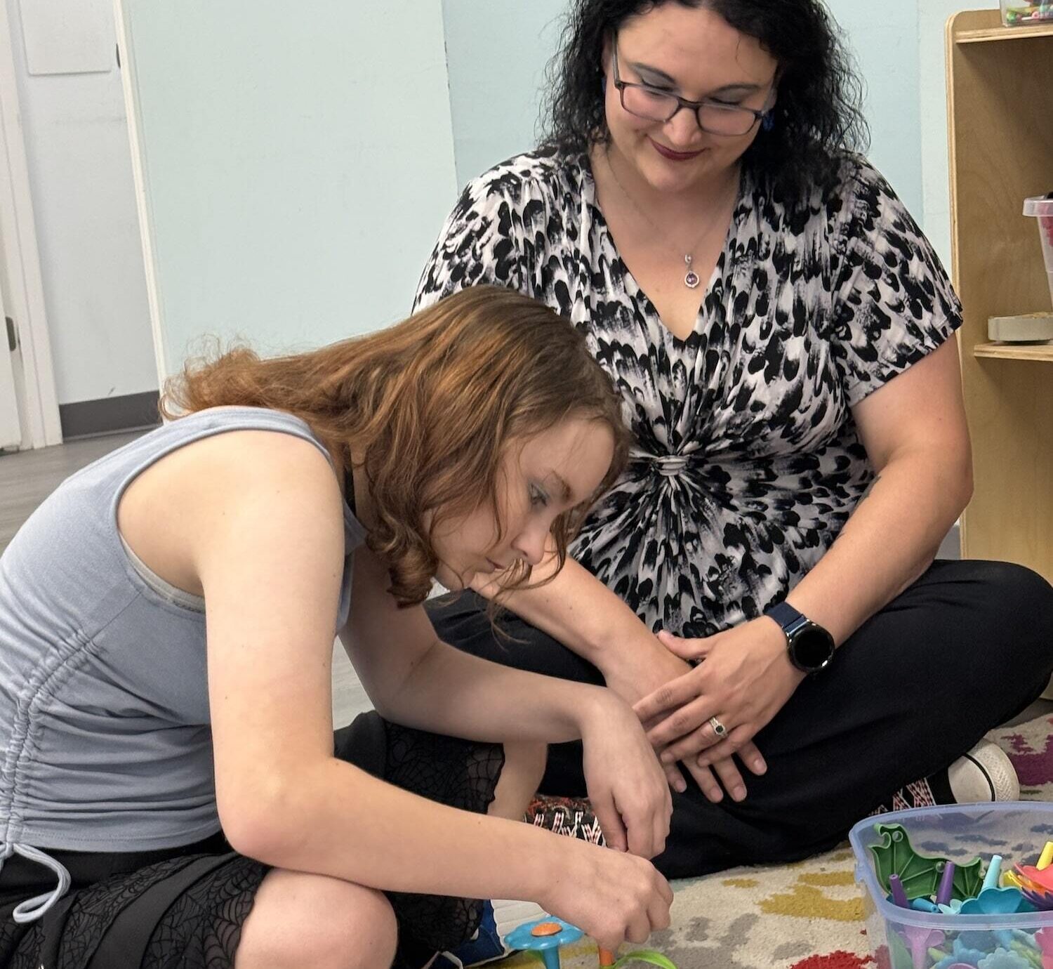 A woman and a girl sitting cross-legged on the floor playing with toys