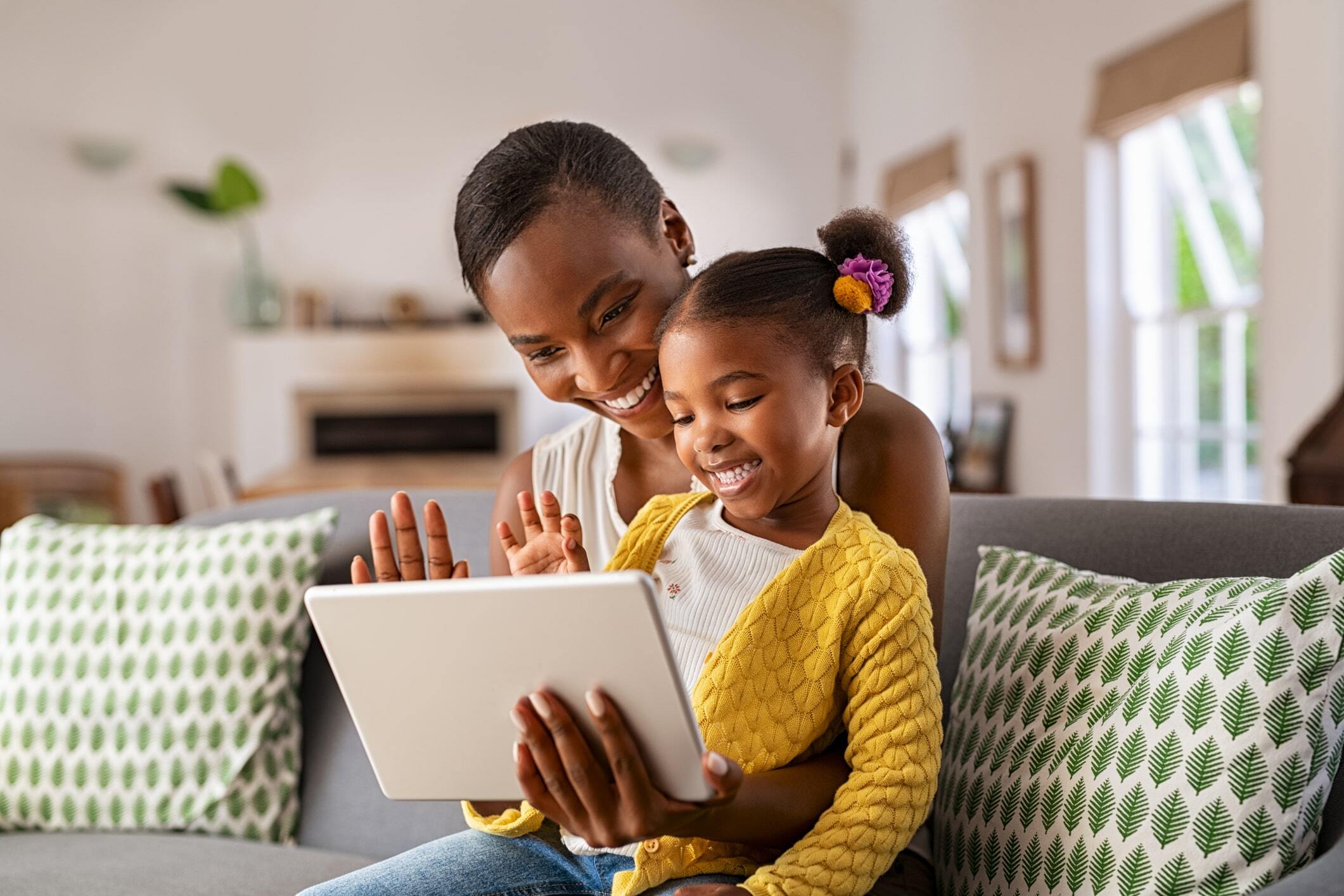 Smiling mother and daughter on a video call using a digital tablet at home, waving hello while sitting on the sofa
