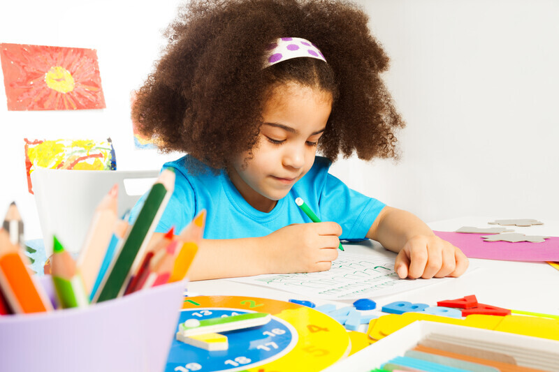 A little girl coloring at a table filled with art supplies, with a colorful clock and a cup of colored pencils in the foreground