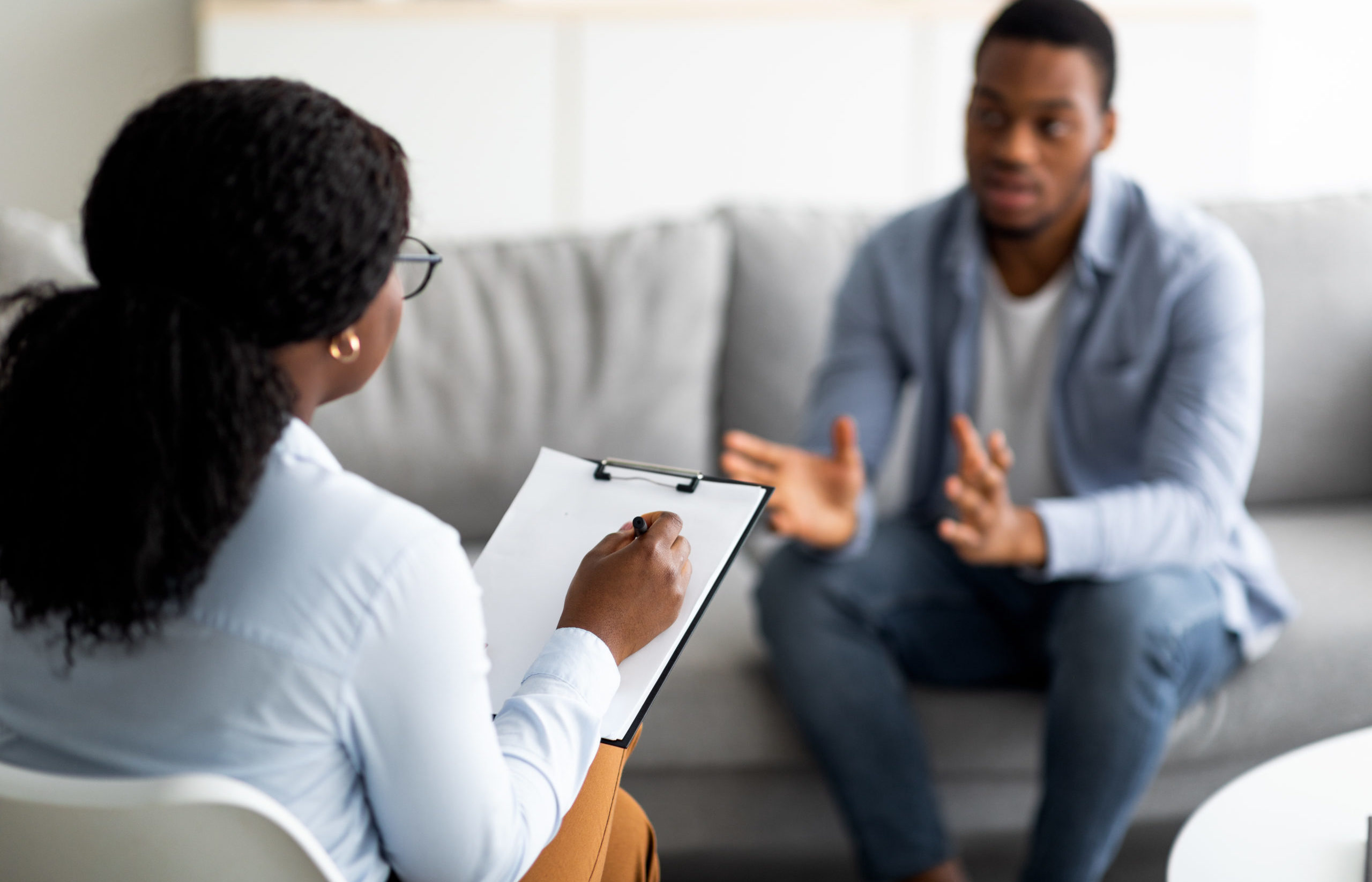 A therapist and a man sit facing one another as the man speaks and gestures with his hands while the therapist takes notes